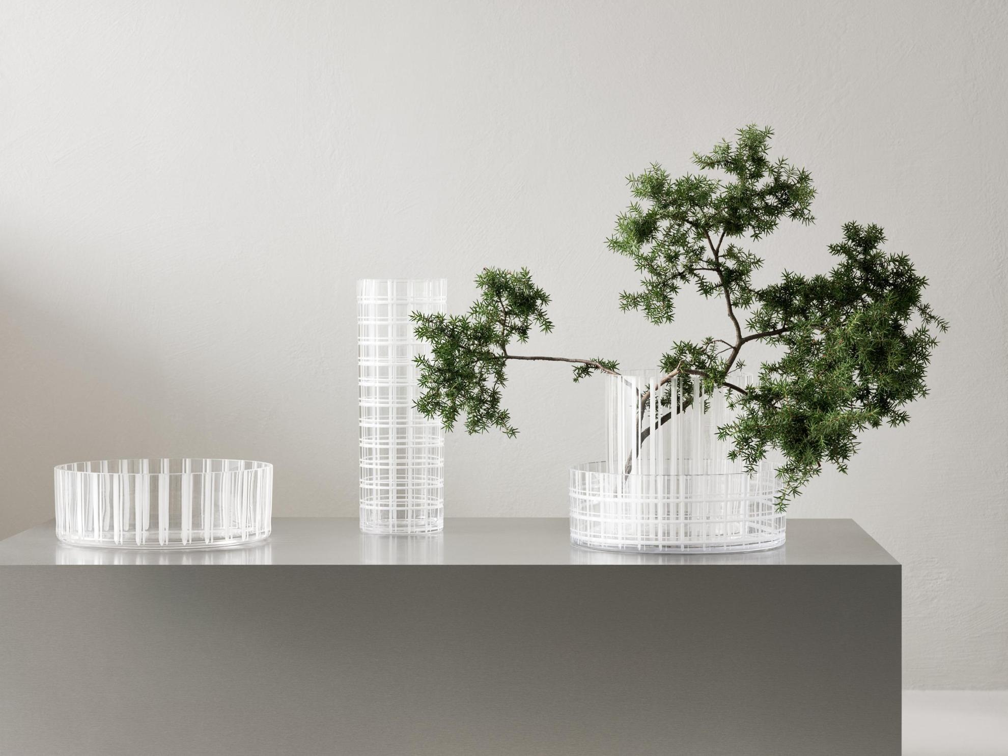 Four transparent vases with white stripes on a grey box. One vase has a small tree branch in it.