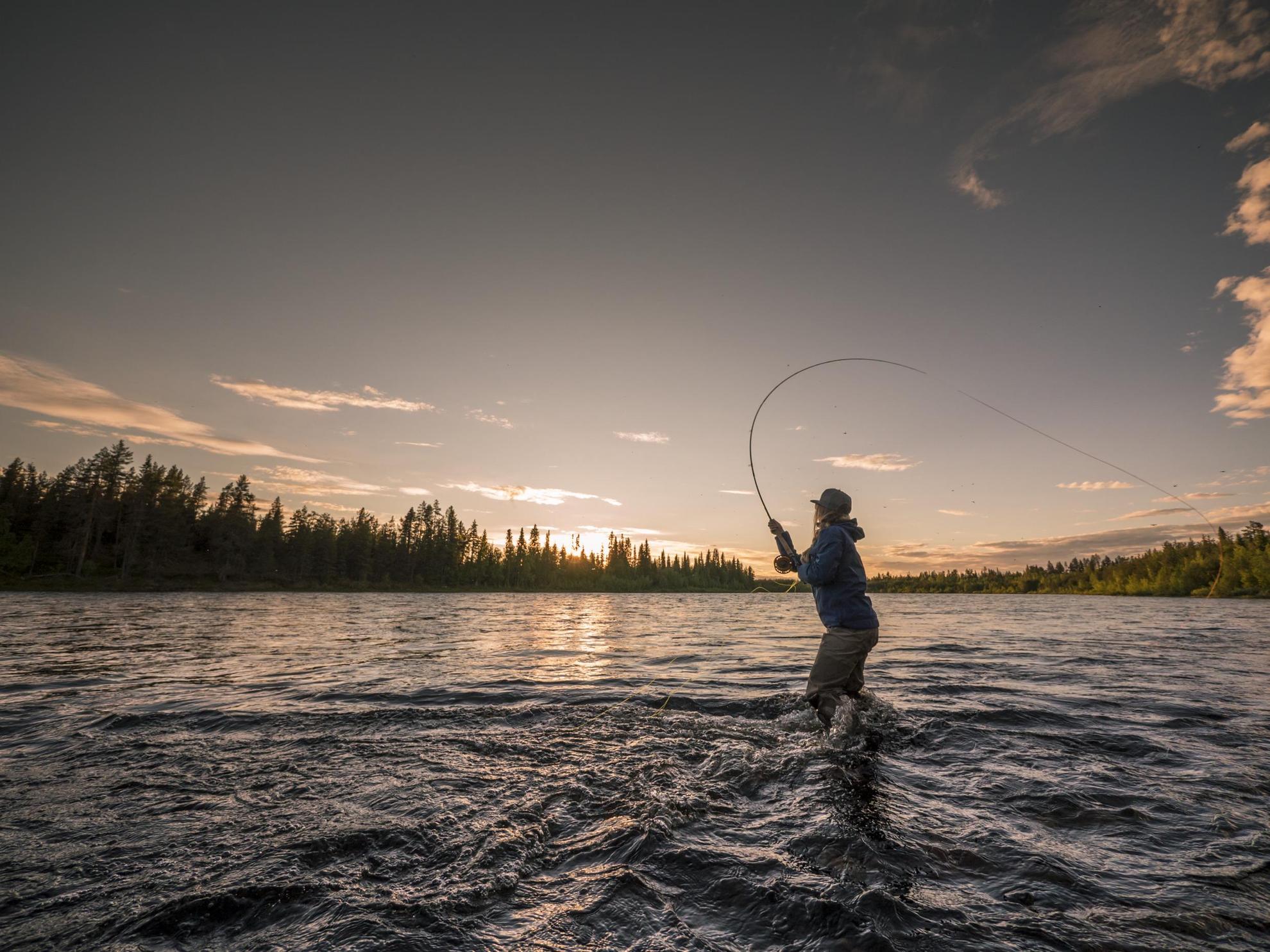 A person is fly-fishing, standing in a river in northern Sweden as the sun is setting.