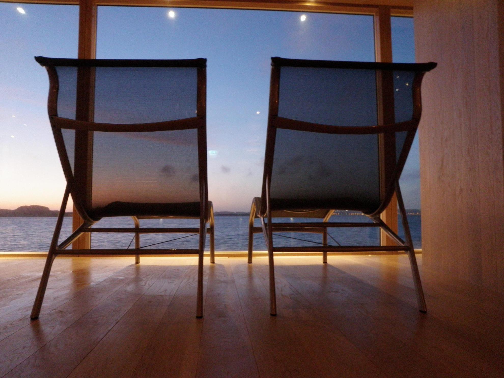 Two empty lounge chairs in front of a window looking out at the ocean by Bohuslän province.