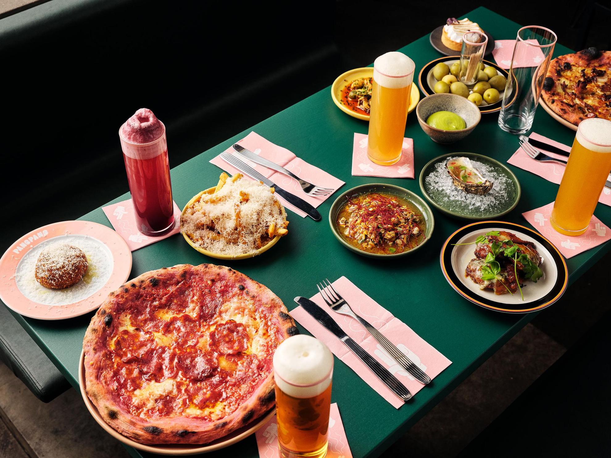 A green table with cutlery, glasses filled with beer and plates with food such as pizza, french fries, oysters and olives.