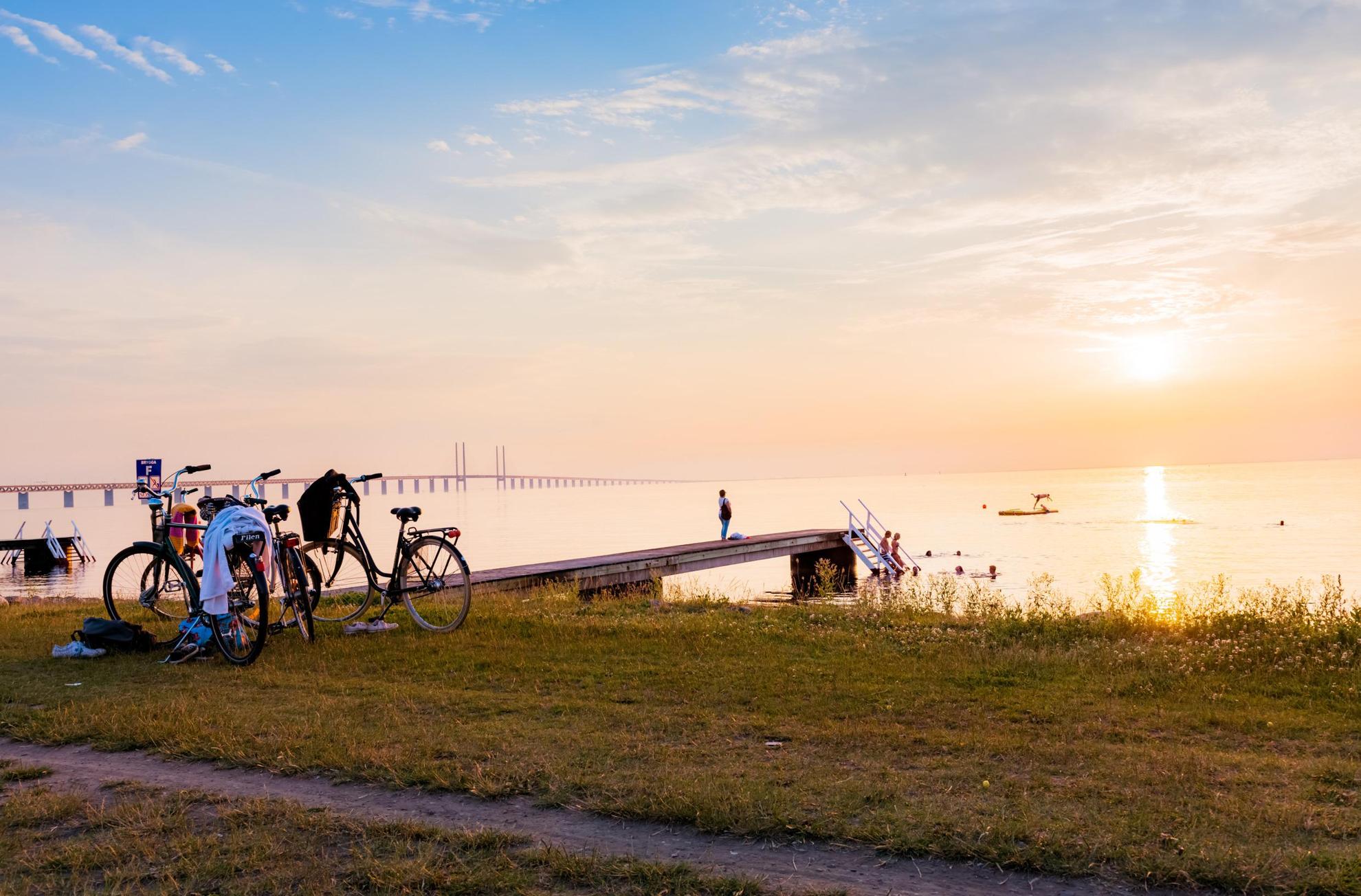 Sunset over a beach with a jetty. Two bikes are parked on the grass above the beach and several people are swimming in the sea. The Öresund Bridge in the background.
