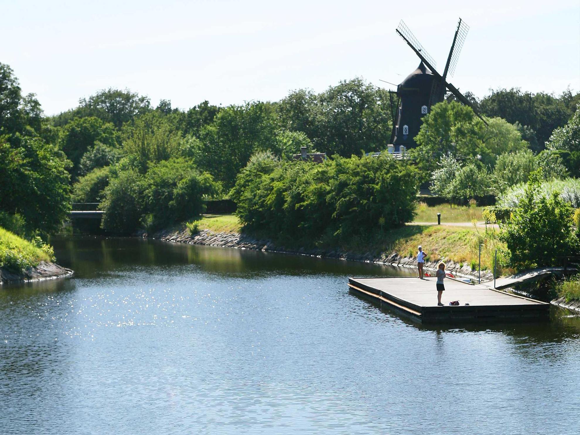 Two women standing on a jetty on a canal in a city garden. There is a windmill surrounded by greenery in the background.