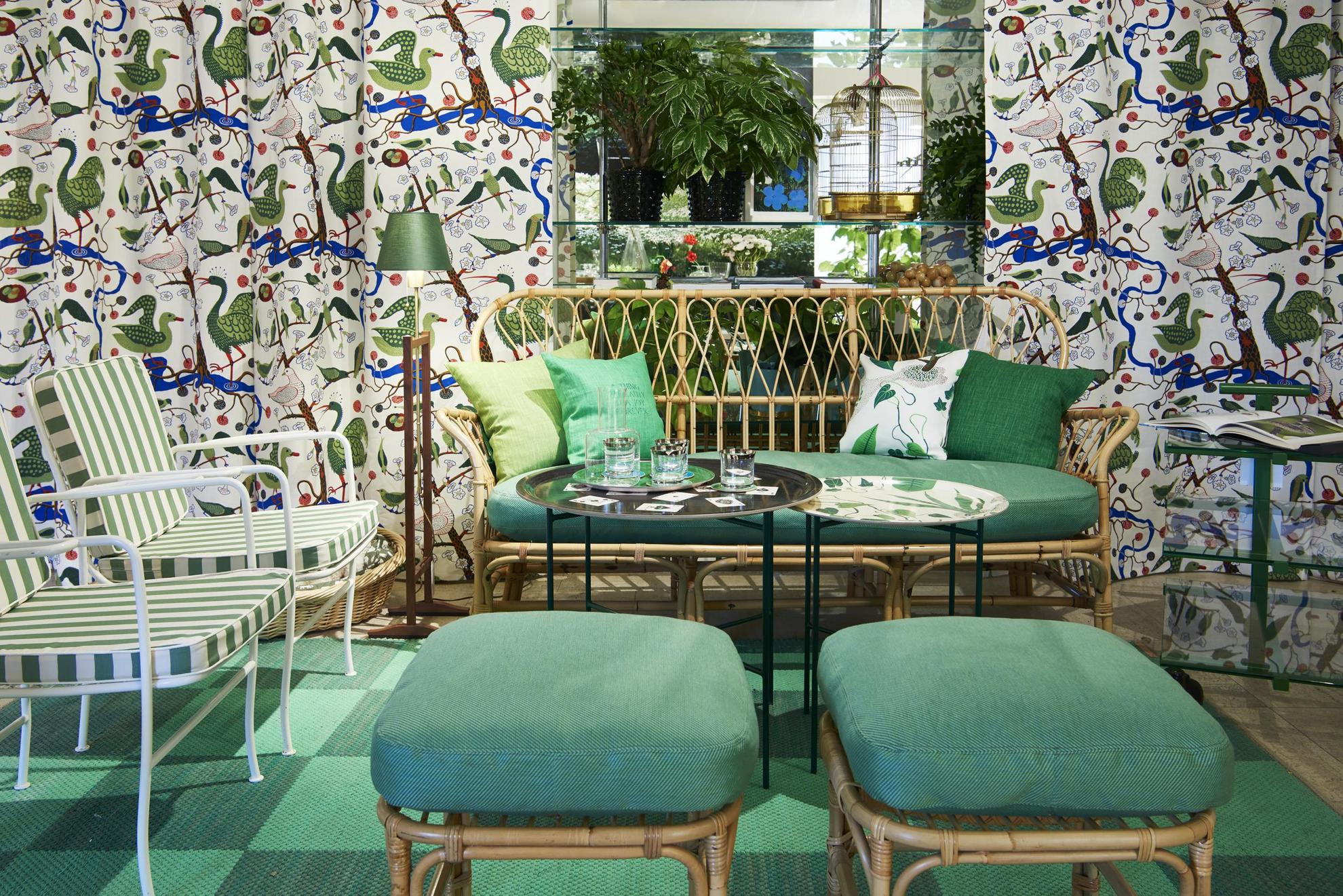 A green environment in a design shop. A sofa, two armchairs and two footstools around a table. Behind the furniture group there are colorful curtains with birds in green.