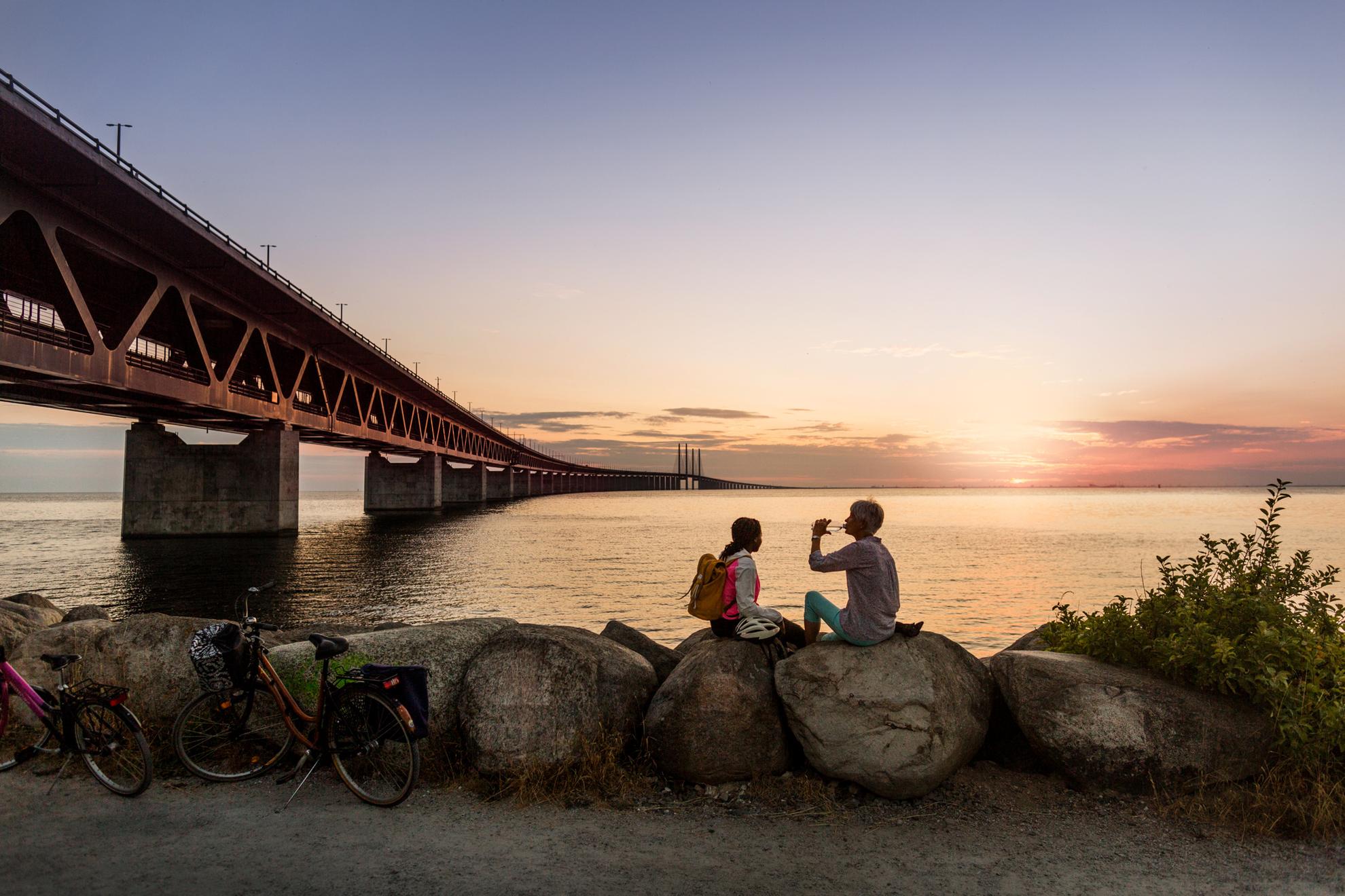 Two women sitting by Öresund Bridge in Malmö at sunset, drinking water from a bottle, two bikes parked on the gravel road.