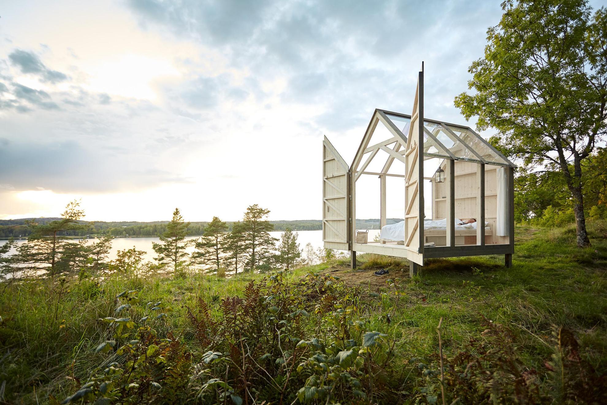 A glass cabin in nature, with doors wide open. Sea and forest in the distance.