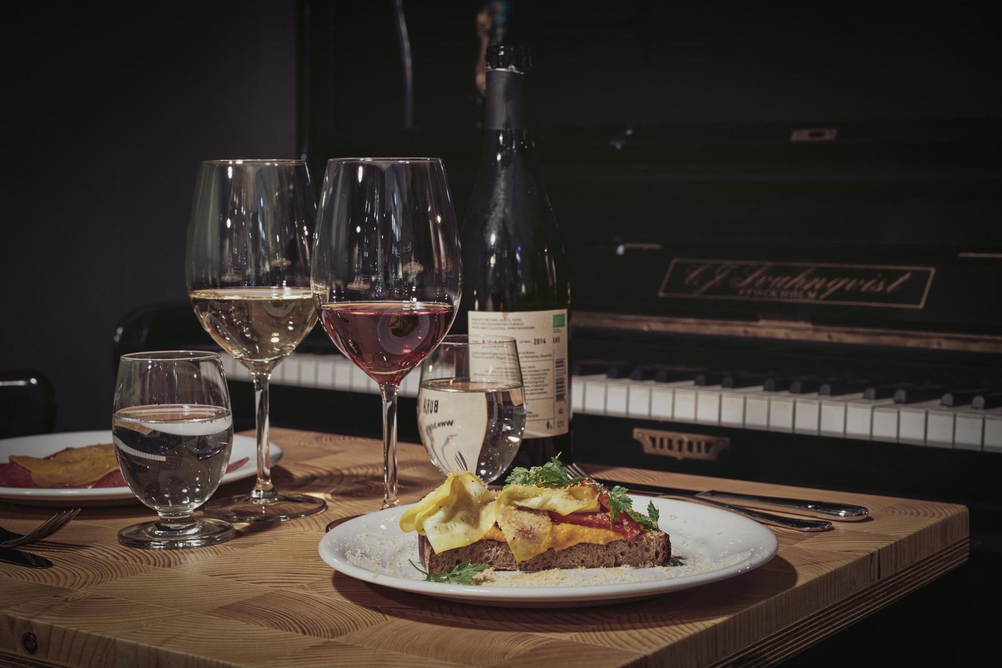 A table set with a wine bottle, two plates of food and two glasses of wine. The table is standing next to a piano.