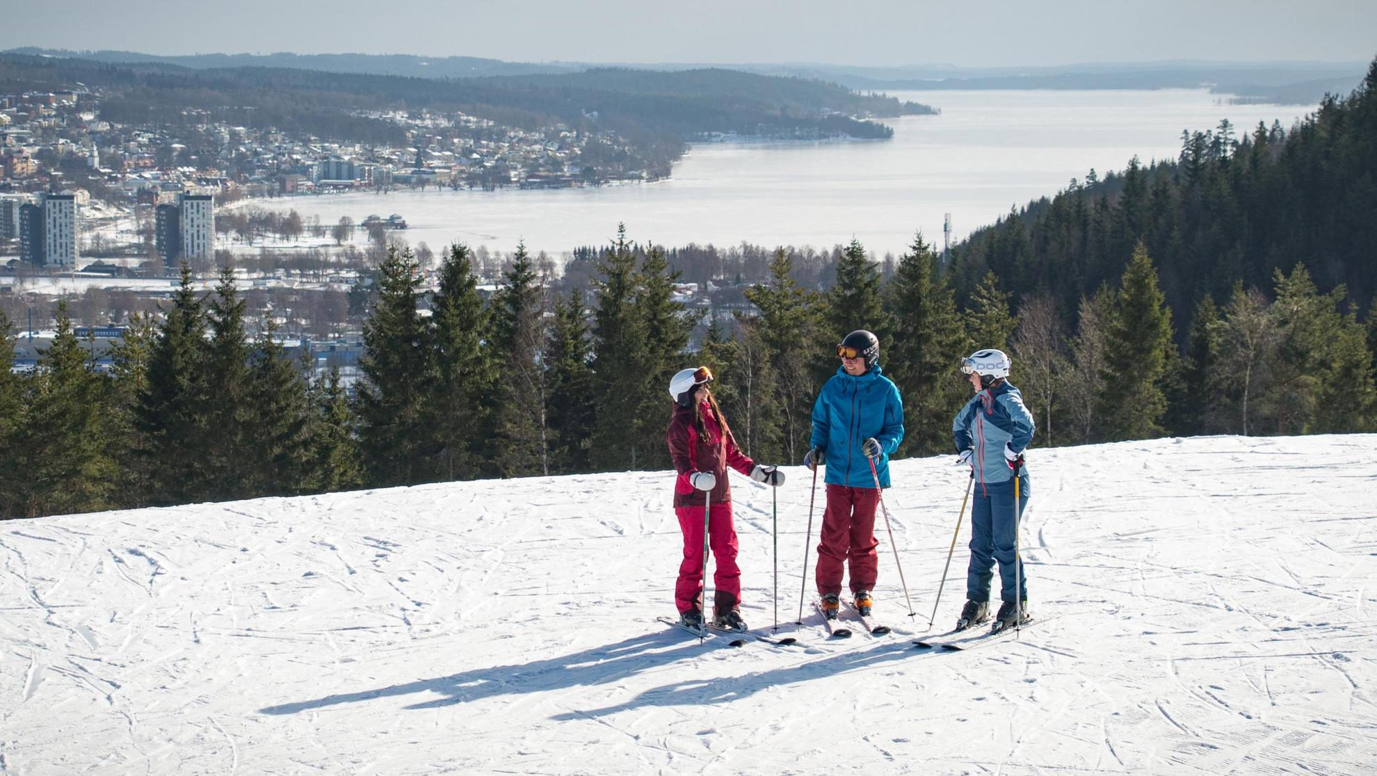 Three people talking with each other, standing in a ski slope. The city of Ulricehamn is in the background.