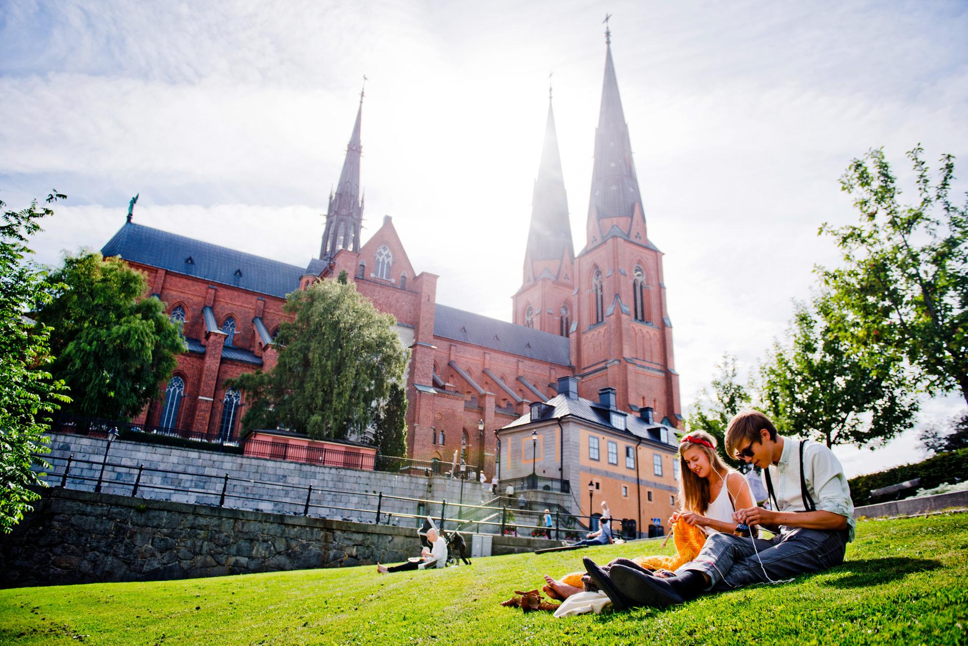 Sunny view over Uppsala Cathedral with people in the foreground sitting on the green grass.