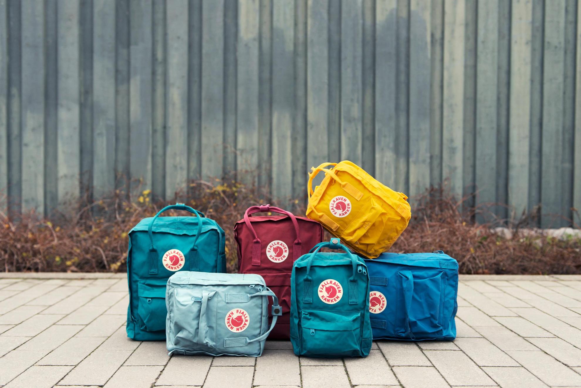 Different-coloured Kånken backpacks arranged on a sidewalk.