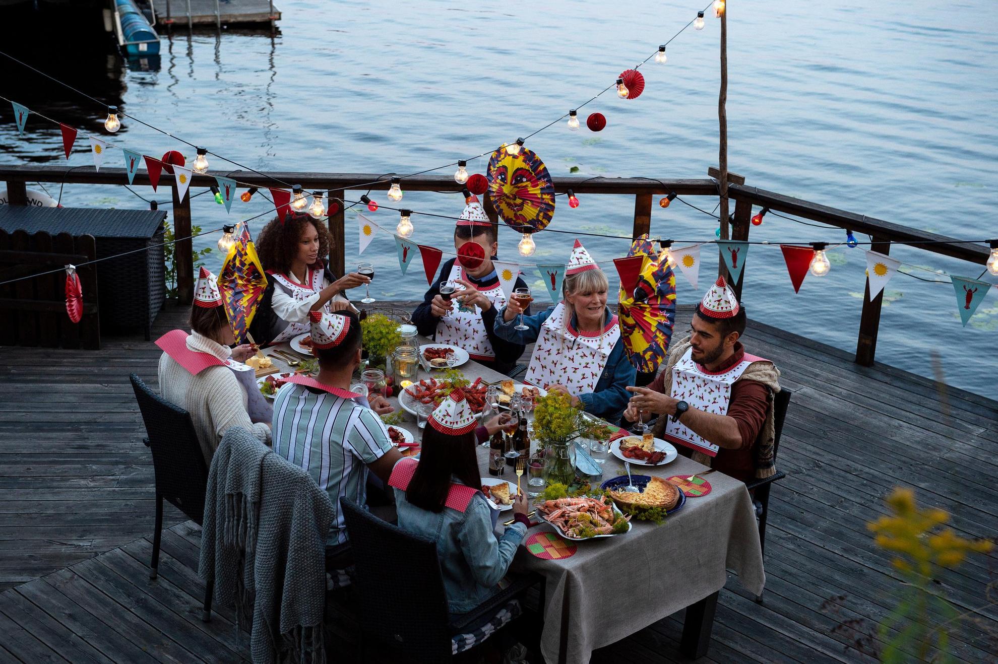Crayfish party at a jetty by the sea. Seven peoples around a table dressed in paper hats and bibs are eating crayfish. There are paper lanterns, lights and decorations above the table.