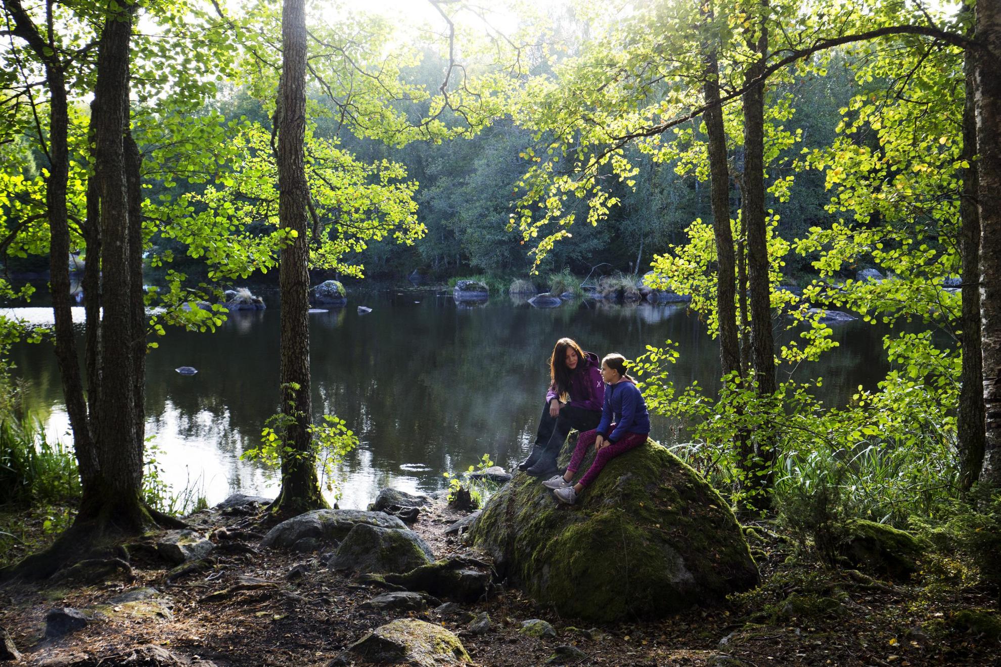 Two girls are sitting on a rock next to a lake in nature.