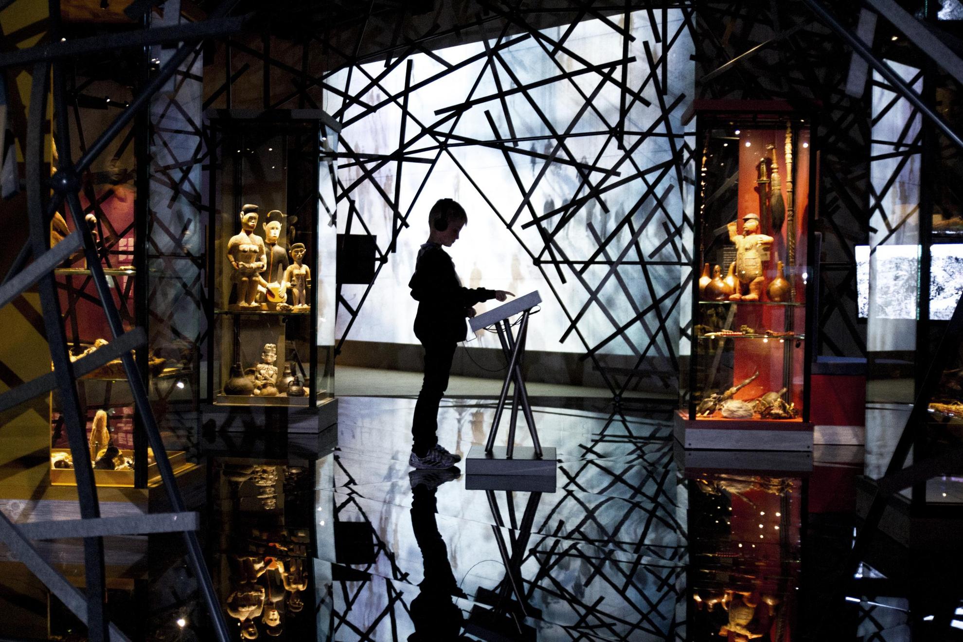 A child is standing in front of a display in the middle of an exhibition room. The room is dark but illuminated by a large screen in the back, surrounded by crossing sticks, which creates a dramatic effect with the visitors silhouette in the middle.
