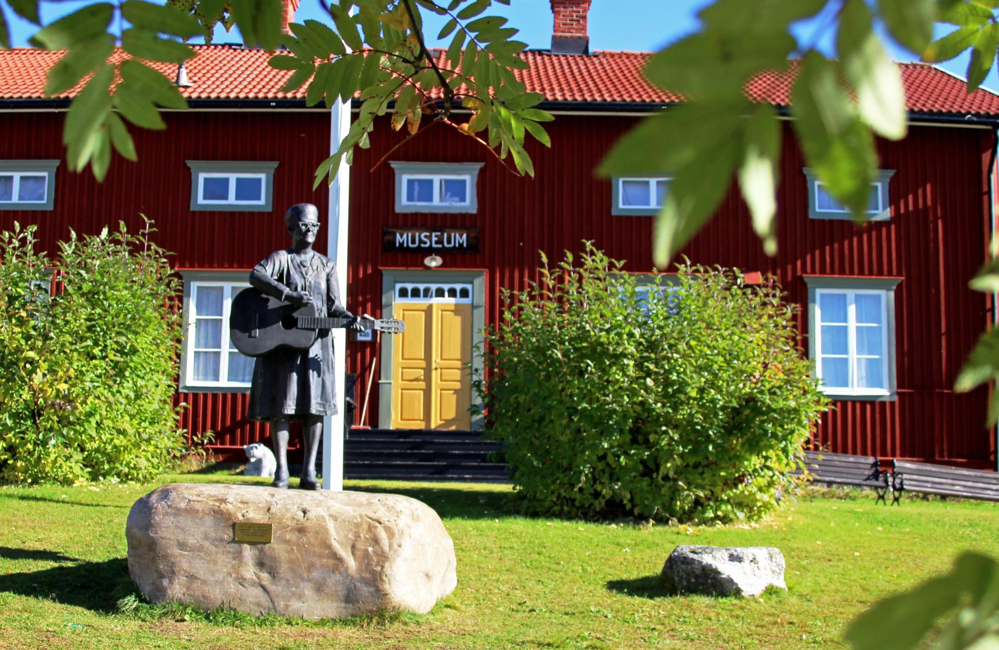 A large red wooden house with a yellow door, surrounded by green bushes. In the foreground is a flagpole and a statue of a woman with a guitar.