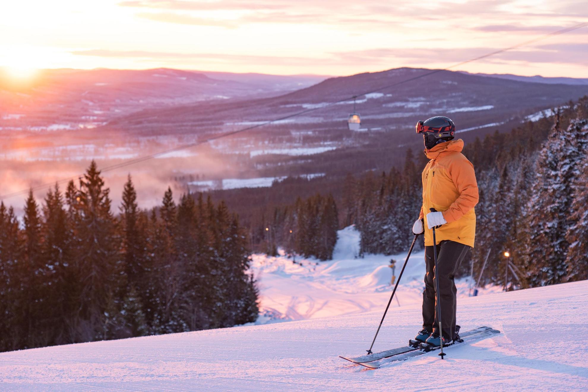 A person with ski gear is standing on a ski slope looking at the sunset.