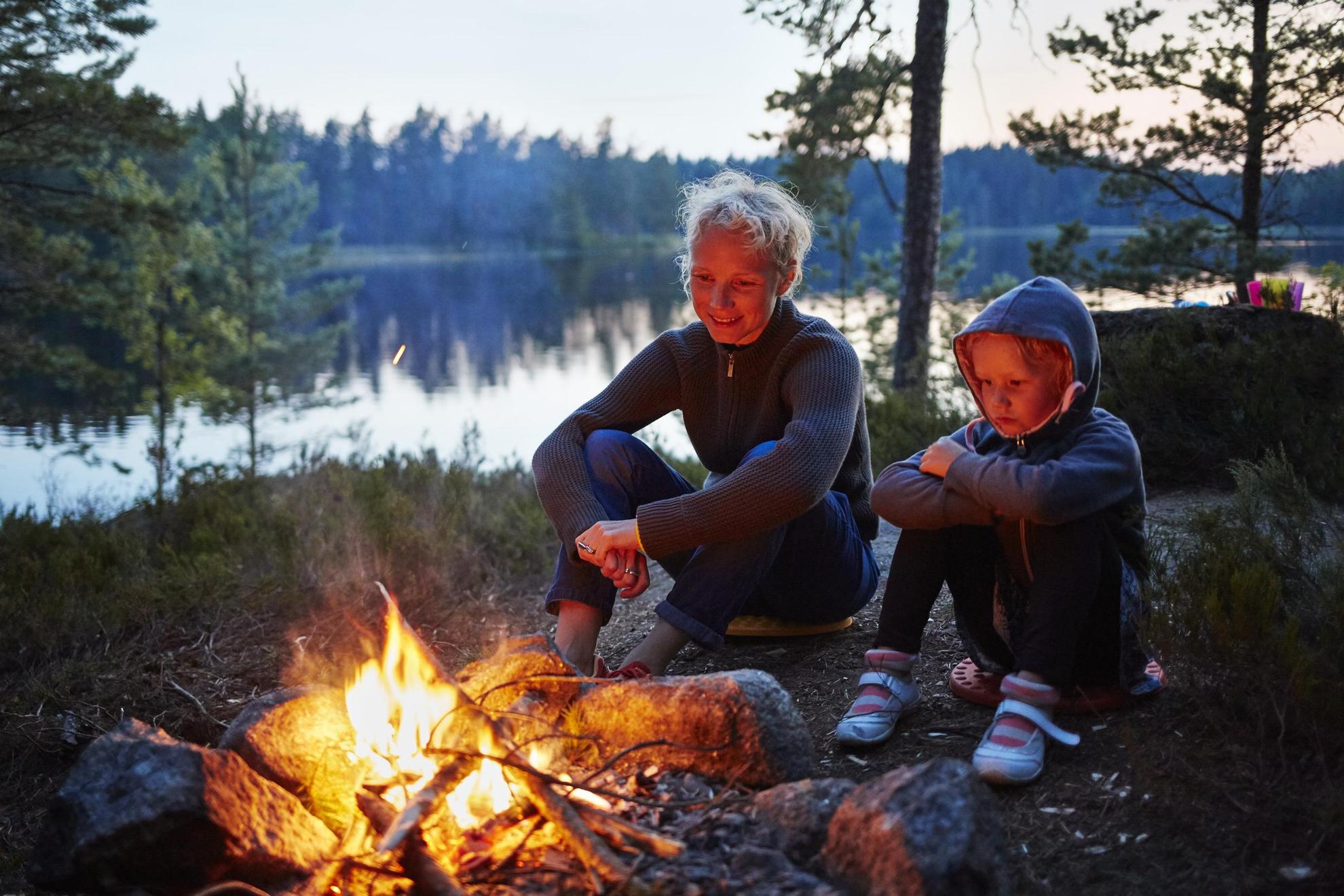 It's evening and two people in Sweden are sitting by a fire in the woods next to a lake.