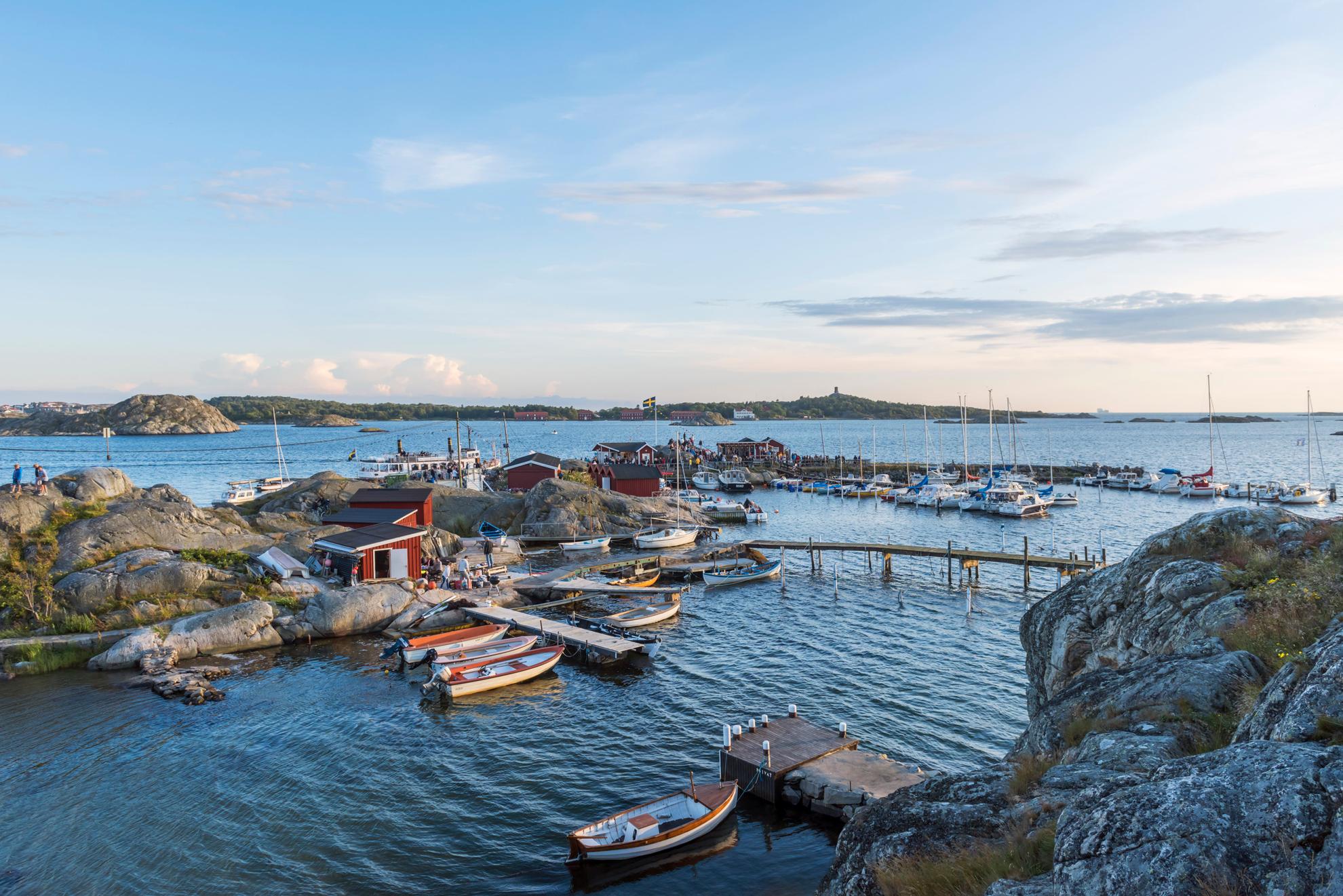 The island of Brännö in the Gothenburg archipelago with boats, rocky cliffs, and piers, red small houses, a Swedish flag, and people in the sun.
