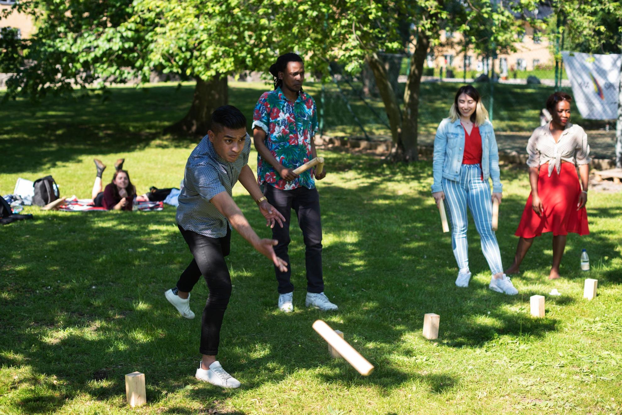 A group of people in a park are throwing sticks at wooden blocks on the grass.