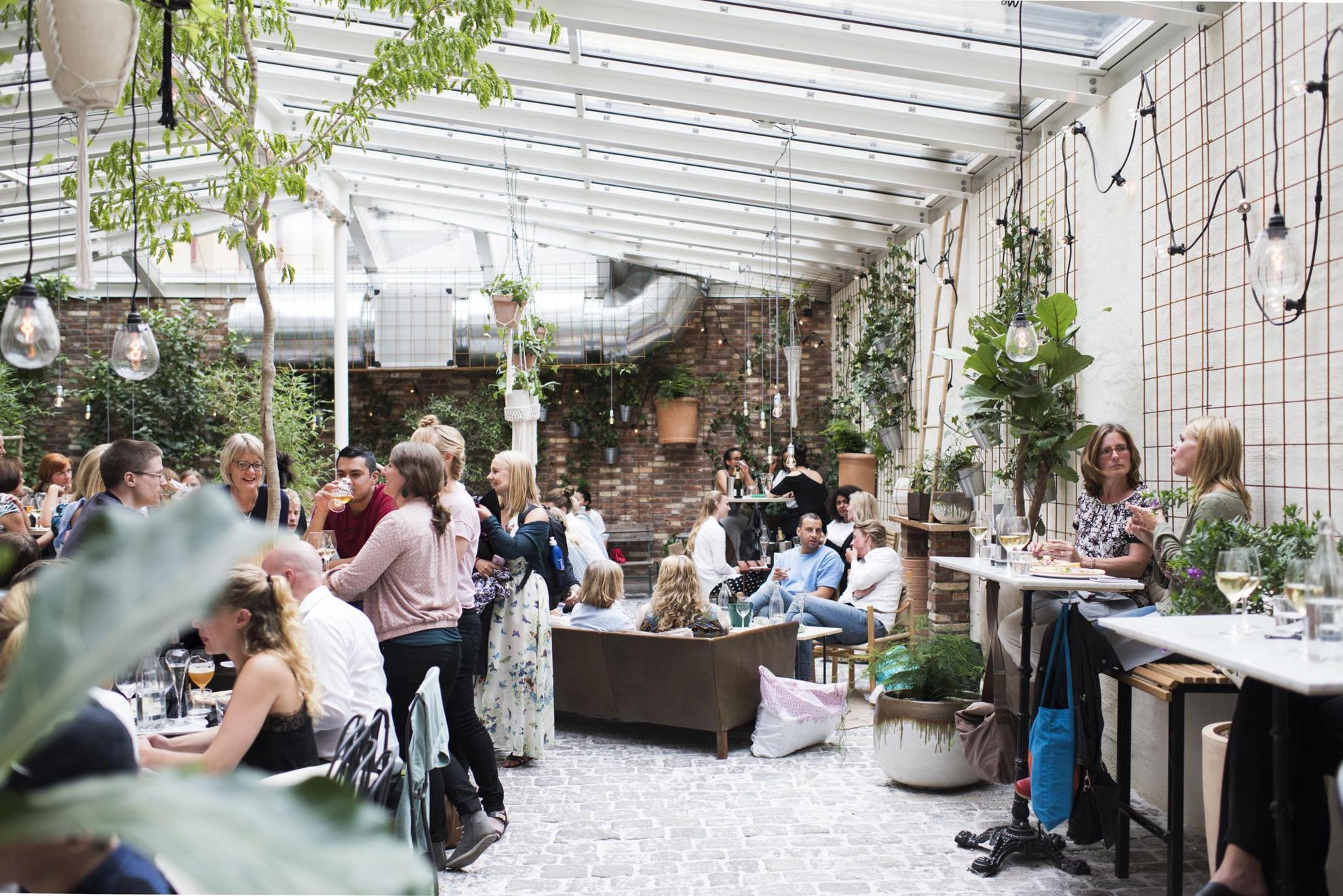 People enjoy drinks and food at a crowded Cafe Magasinet in Gothenburg. The interior is stone floor, brick walls, a glass ceiling and the space is full of green plants.