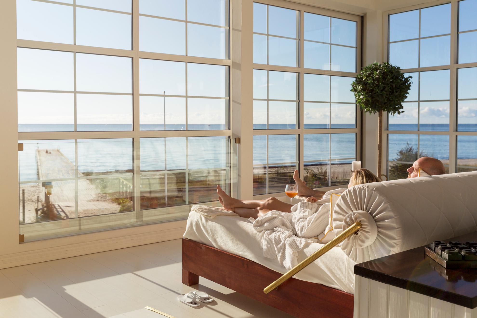 A couple are lying in a large bed next to large windows facing a beach and the sea.