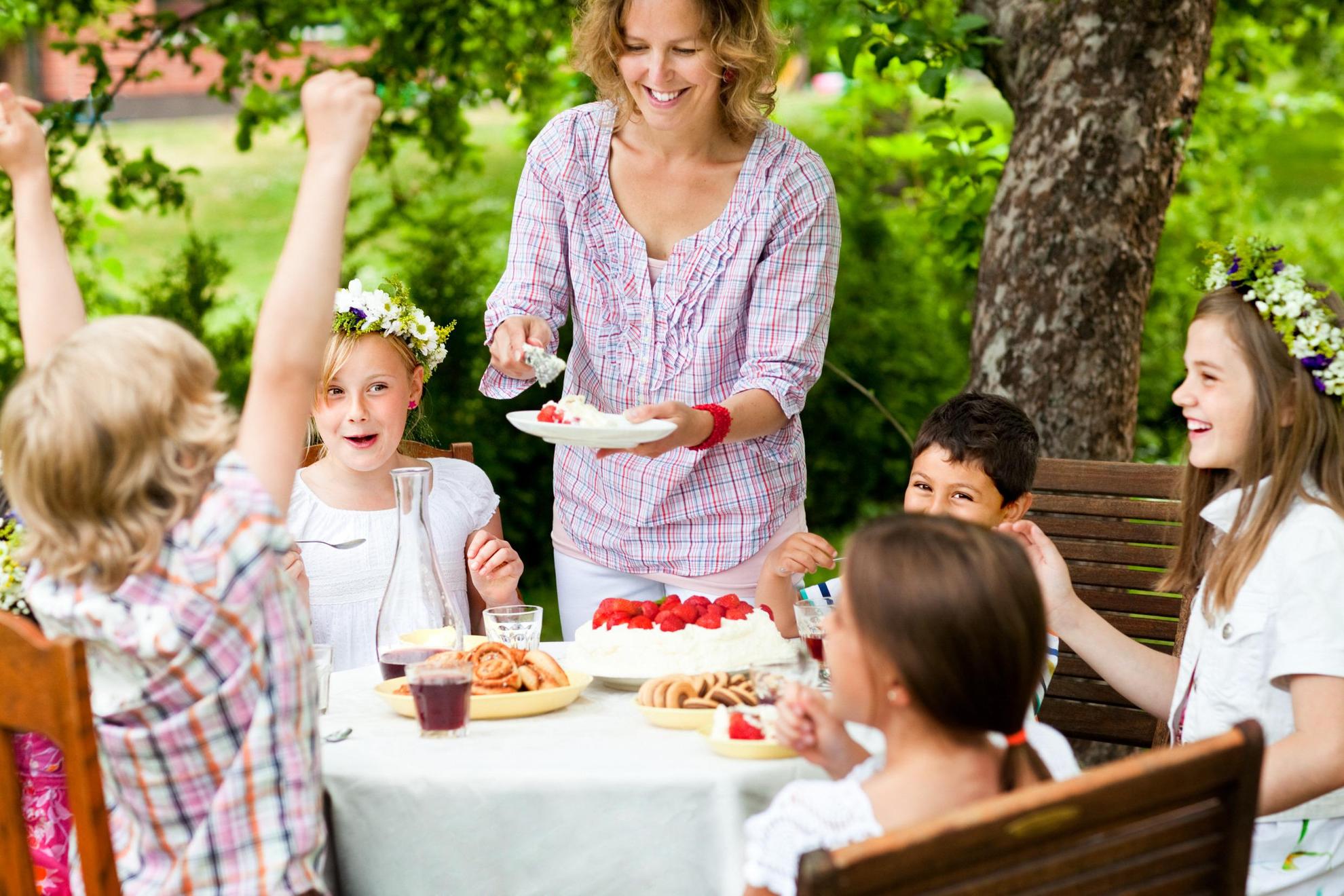 A woman is serving strawberry cake to some children sitting at an outside table. Some of the kids have Midsummer flower wreaths in their hair.