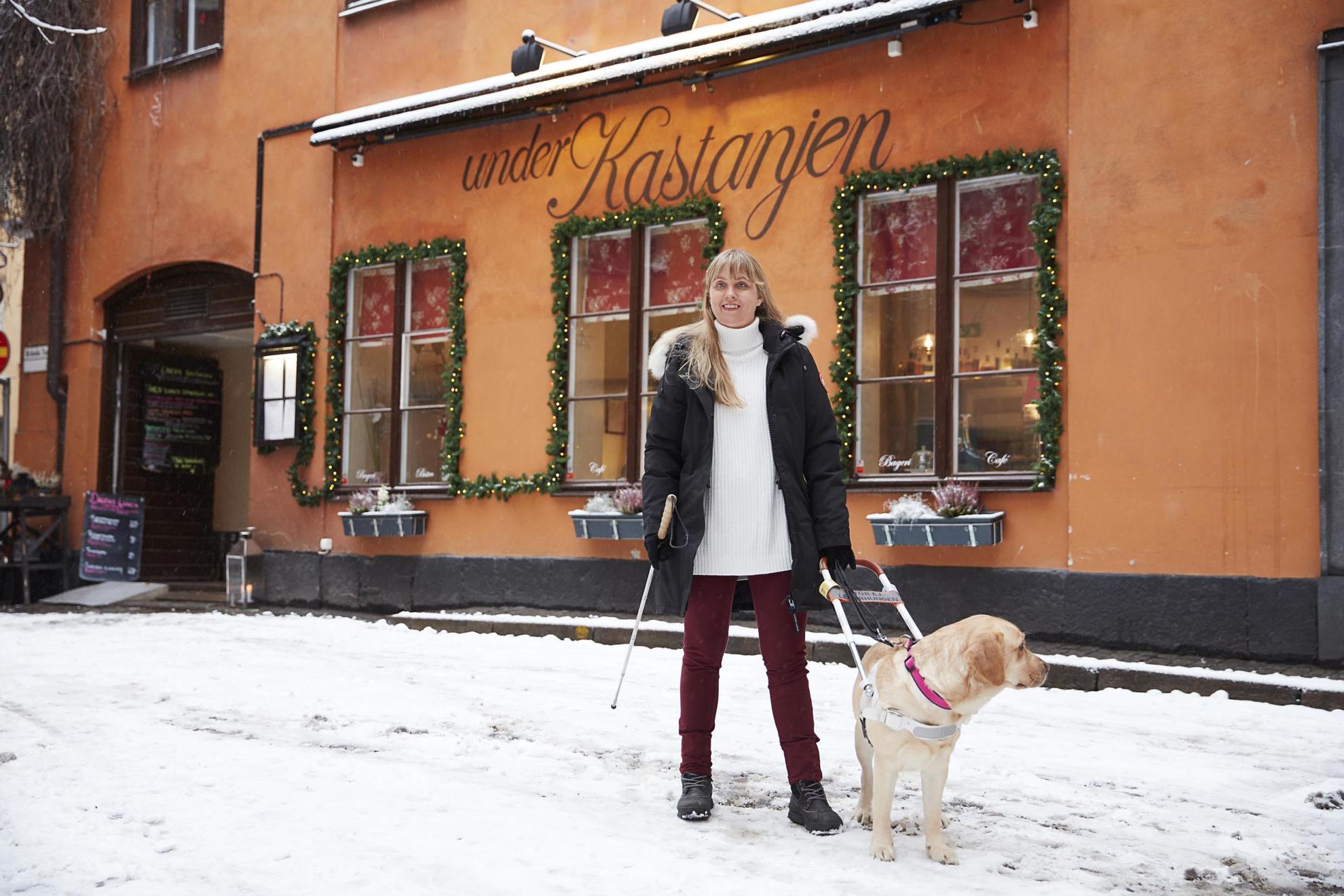 Journalist Anna Bergholtz is standing with her guide dog outside a restaurant in Stockholm old town. There is snow on the ground.