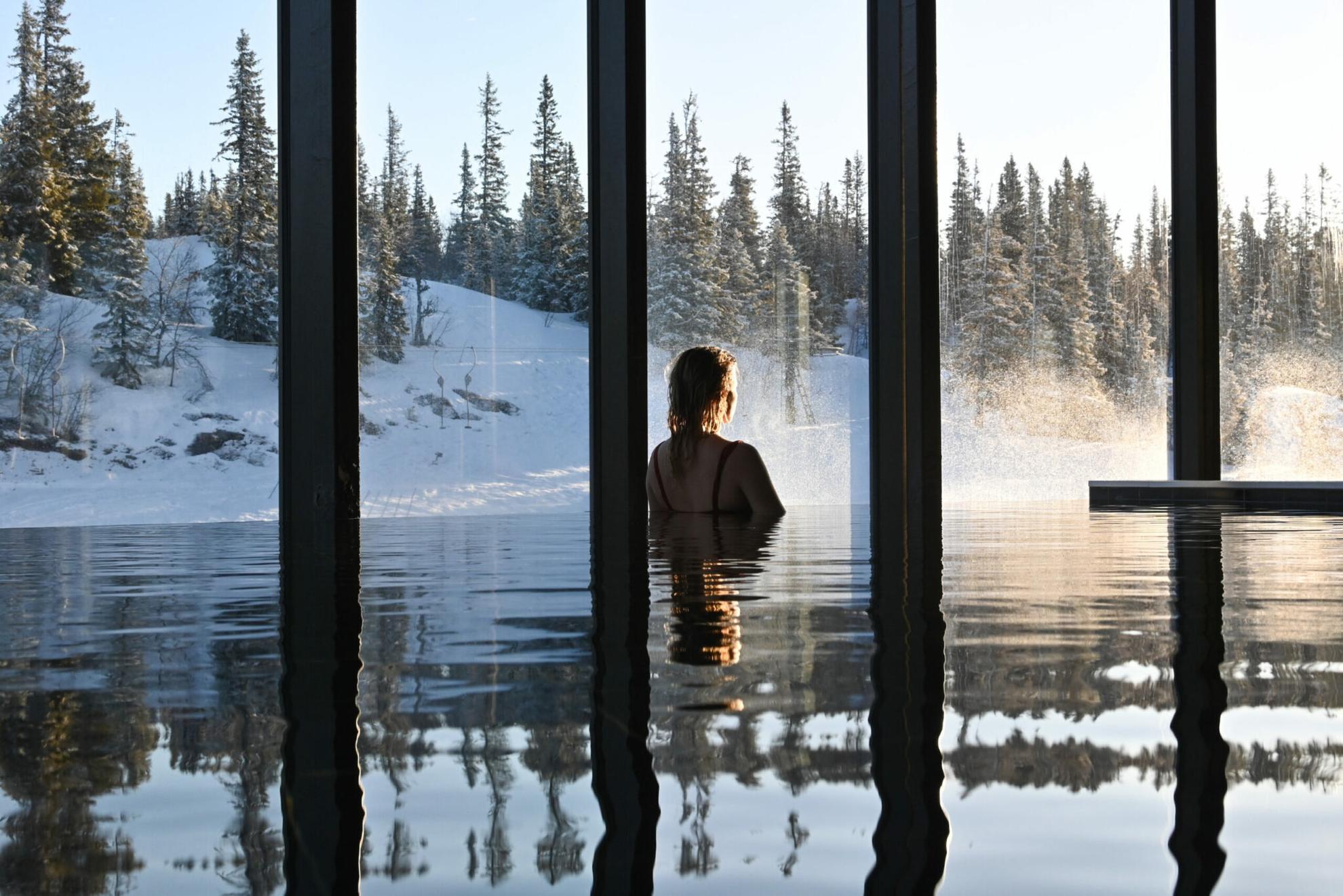 A person in an indoor infinity pool looking out through the big windows to the snow-covered forest.
