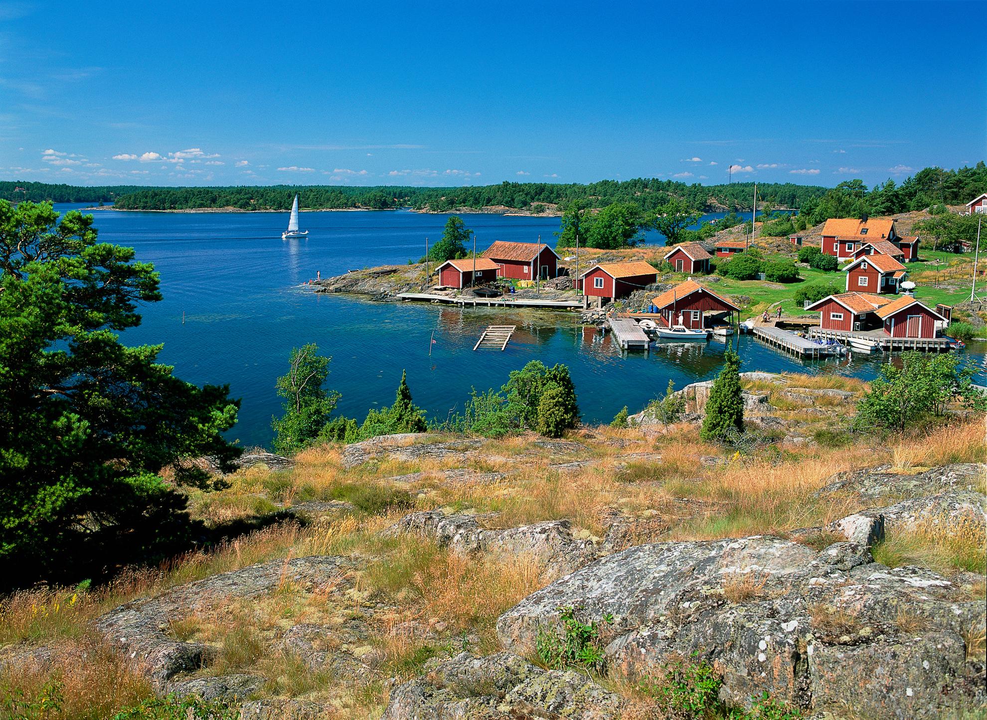 Red cottages on an island in the archipelago on a sunny day. A sailboat sails by the islands.