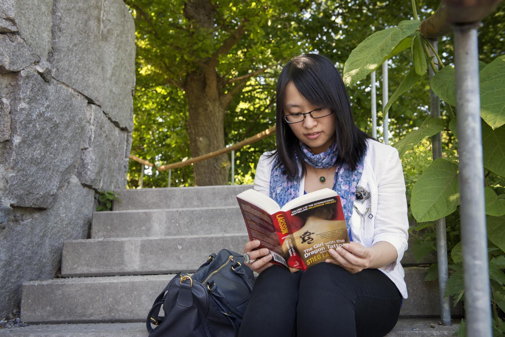A woman is sitting on stone steps outdoors, reading a Stieg Larsson book in English.