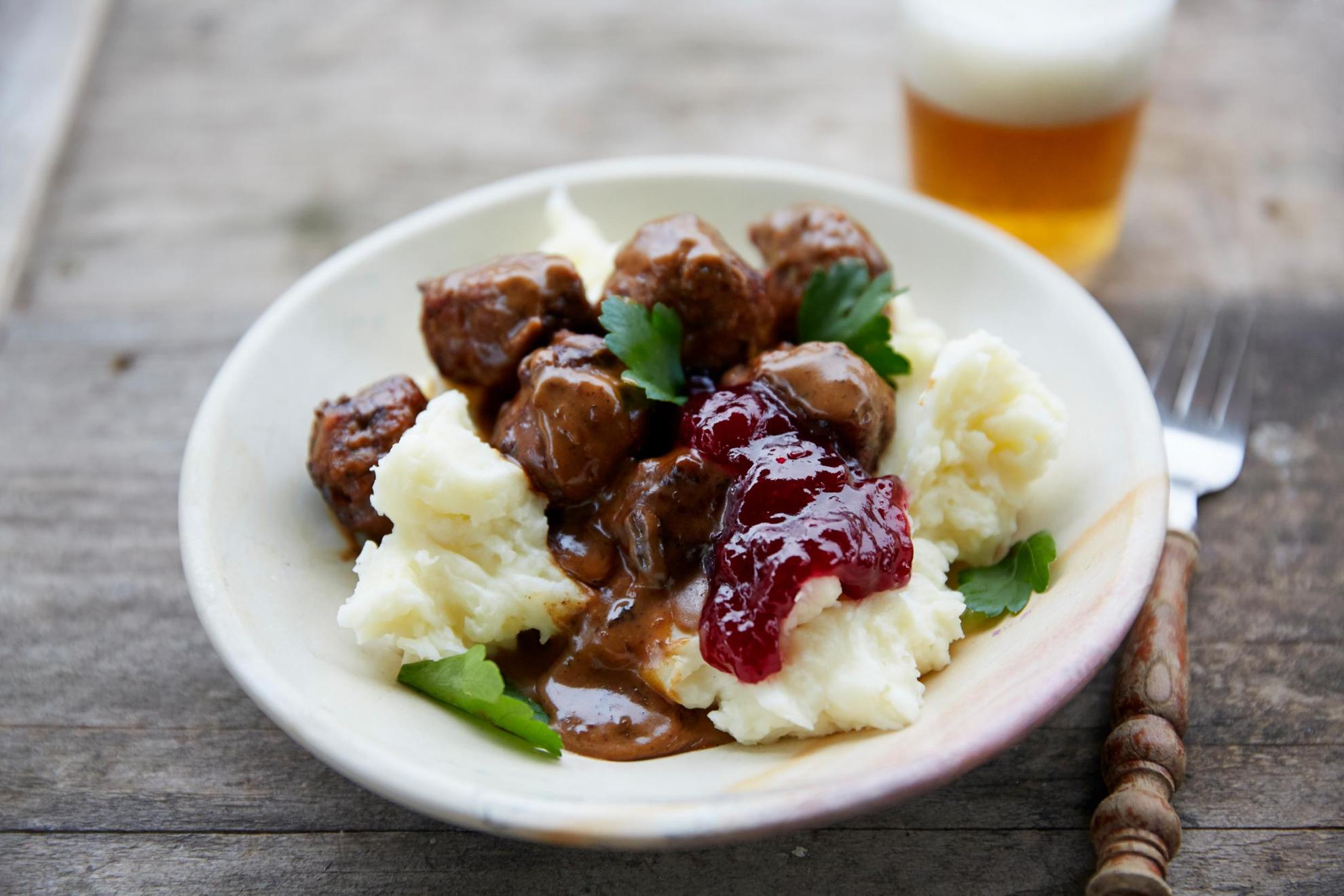 A wooden table is set with a glass of beer, a fork and a plate of meatballs with mashed potatoes, lingonberry jam and brown gravy.