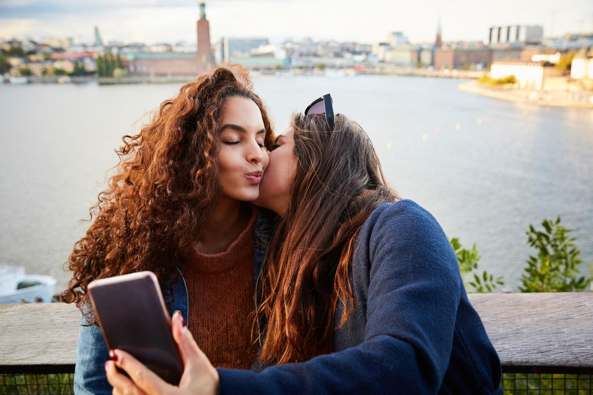 Two women stand on a bridge, taking a selfie while kissing.