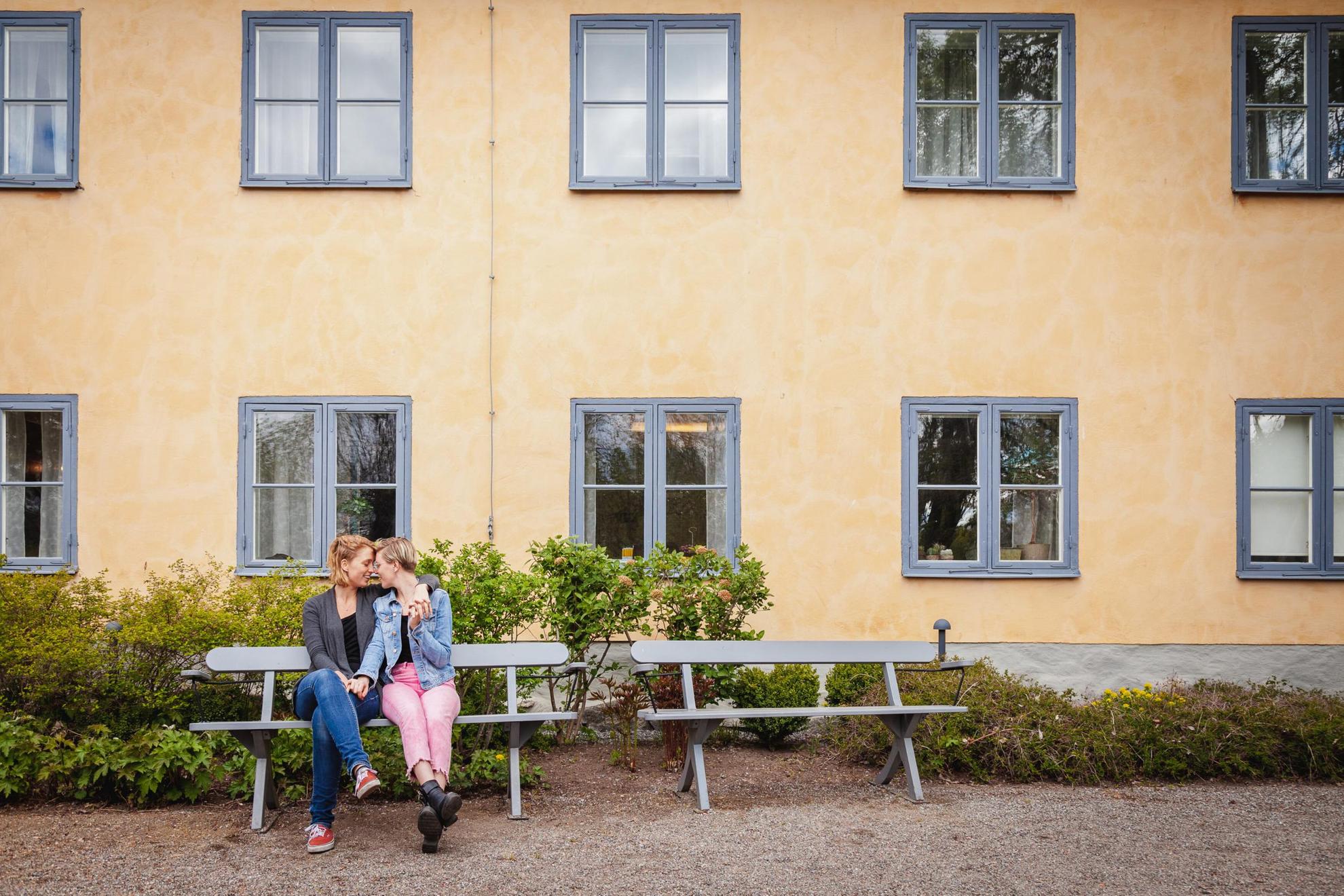 Two women kissing and holding hands while sitting on a park bench outside a house.