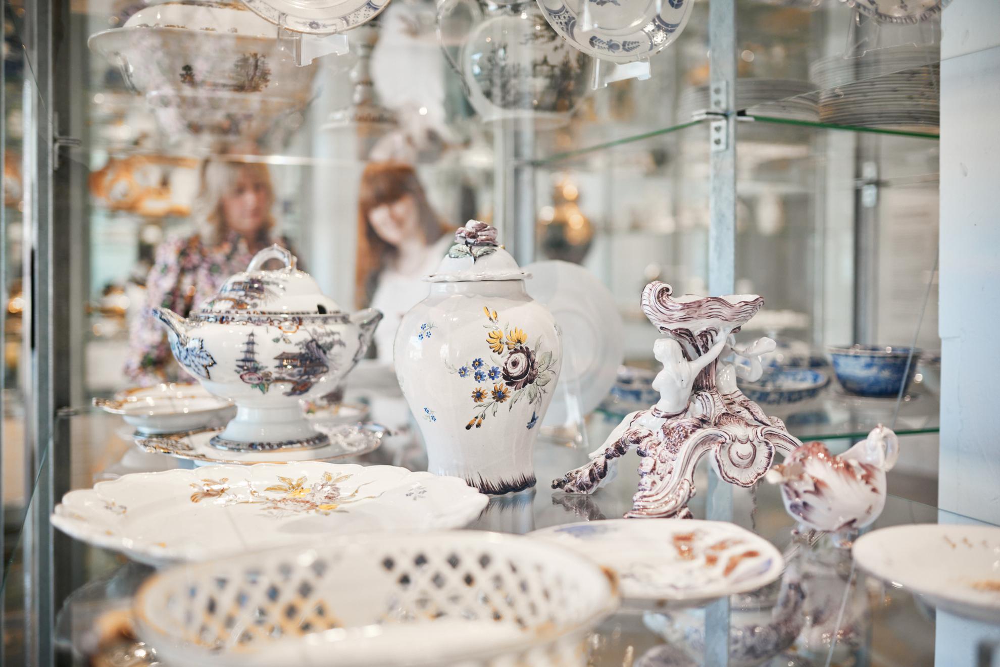 A porcelain teapot, a porcelain bowl with lid and a porcelain ornament, all placed inside a glass stand.