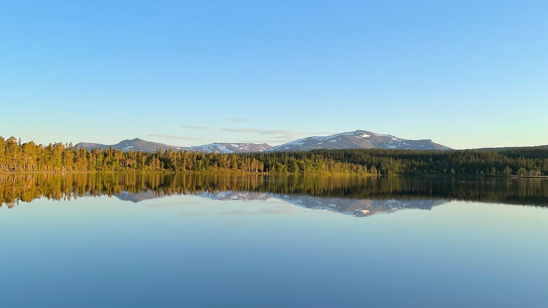The water in Lake Nulltjärn is calm and reflects the blue sky and the surrounding forest and mountain.